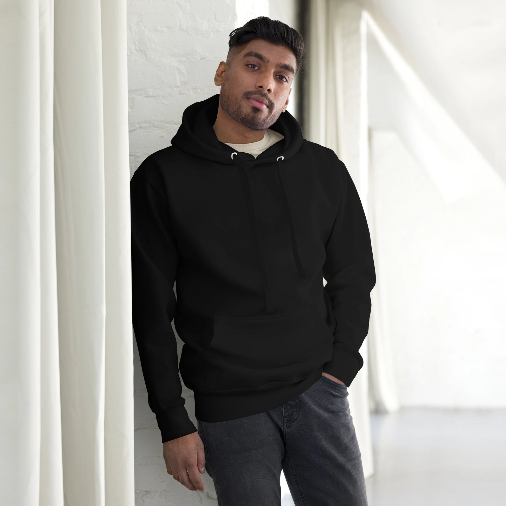 Man standing by window wearing black hoodie, plain front, no visible design.