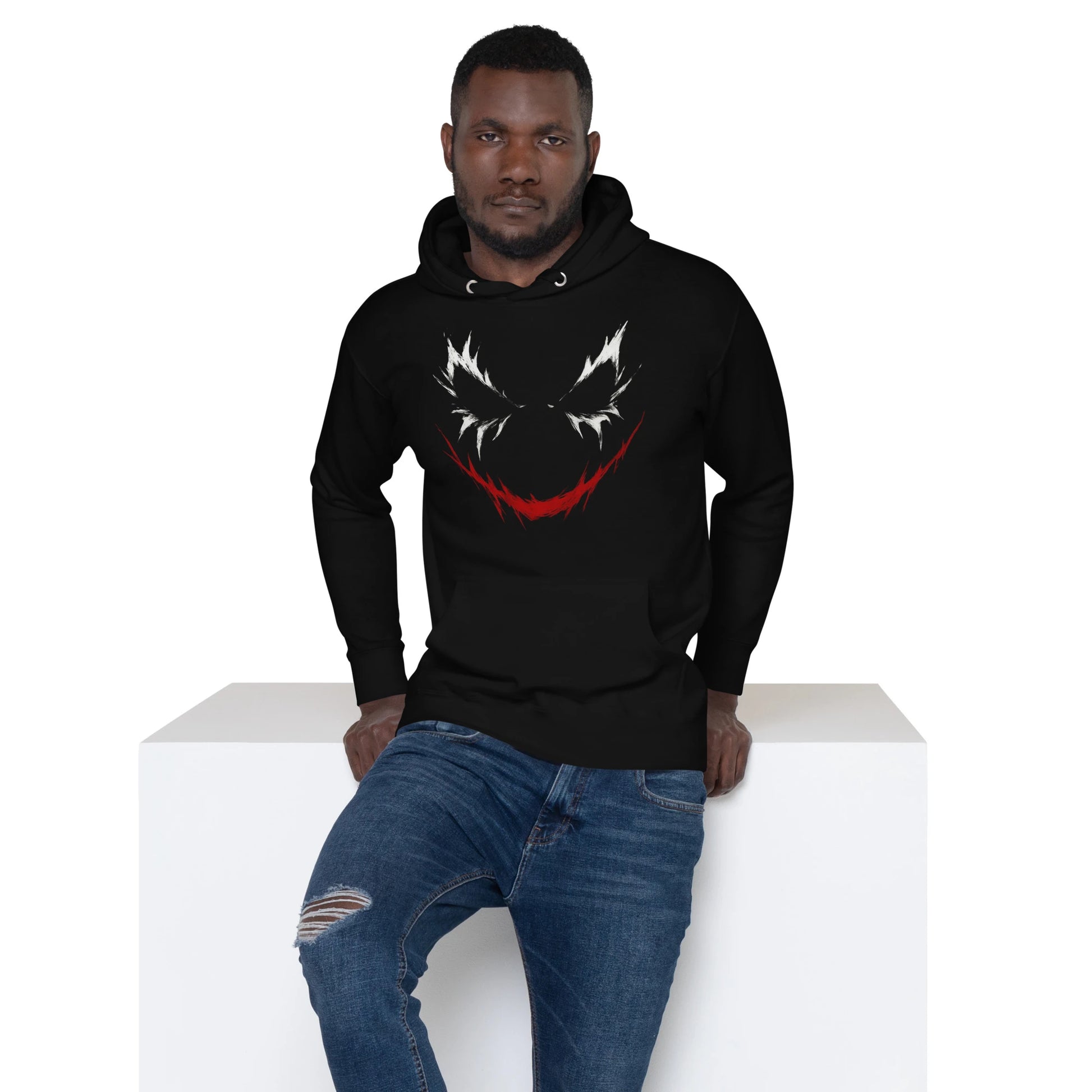 Man seated on white cube, wearing Joker grin black hoodie with intense white and red graphic on the front.