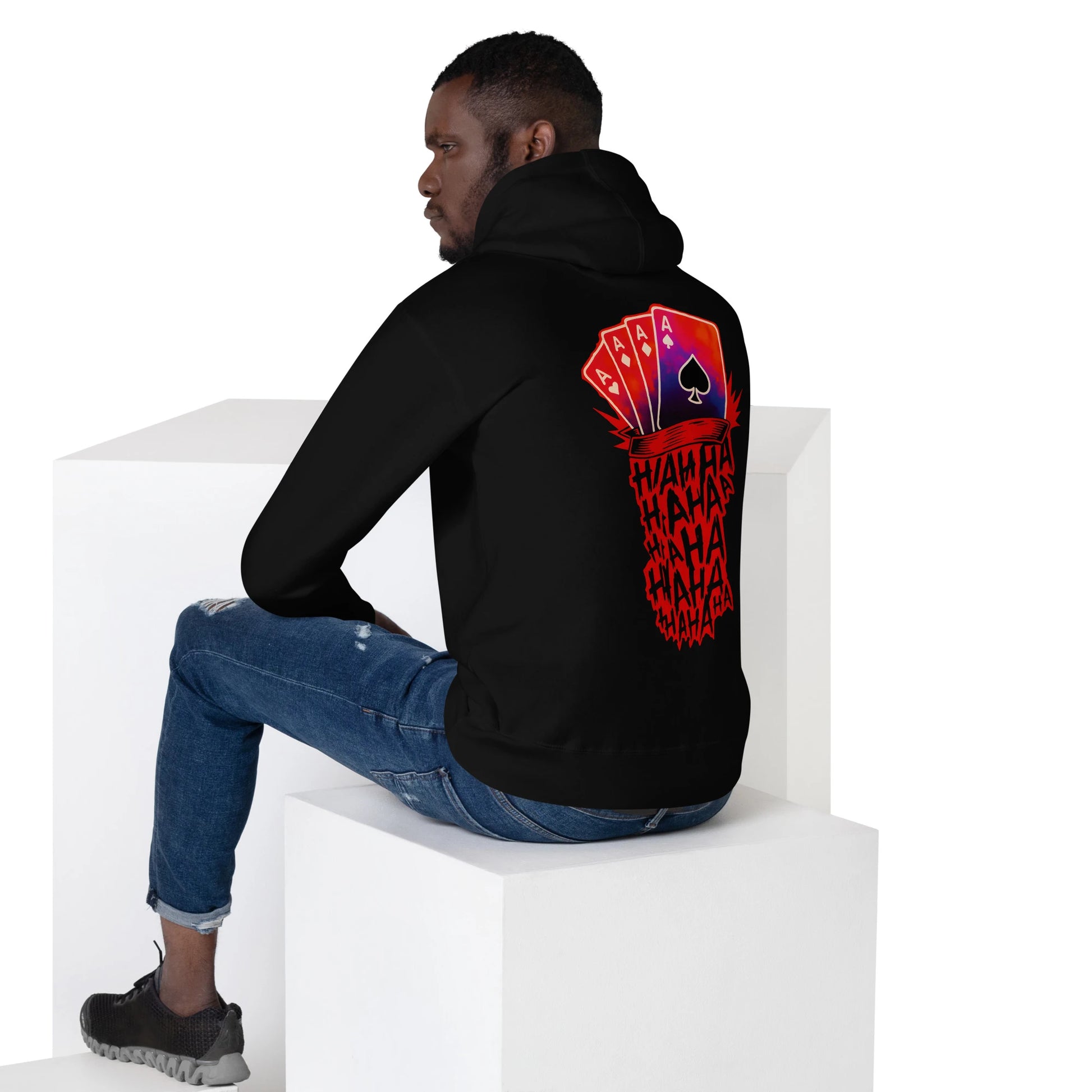 Man seated in studio wearing black hoodie with bold Joker card hand and red 'HAHA' graphic on the back.